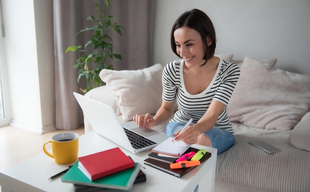 Schöne lächelnde junge moderne Frau, die zu Hause an ihrem Laptop arbeitet, während sie auf der Couch sitzt. Freiberufler, Geschäft zu Hause. Quarantänearbeit