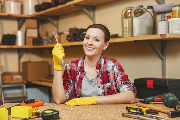 Schöne lächelnde kaukasische junge braunhaarige Frau in kariertem Hemd, grauem T-Shirt, gelben Handschuhen mit Schraubenschlüssel, die in der Tischlerei am Holztischplatz mit verschiedenen Werkzeugen arbeiten. Geschlechtergleichheit.