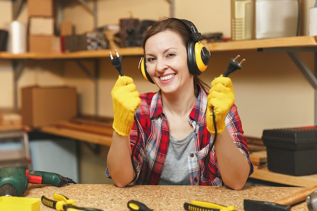 Schöne lächelnde kaukasische junge Frau im karierten Hemd, geräuschisolierte Kopfhörer, die in der Tischlerei am Tisch arbeiten, mit zwei Drähten von Elektrogeräten wie Bohrmaschine und Säge.