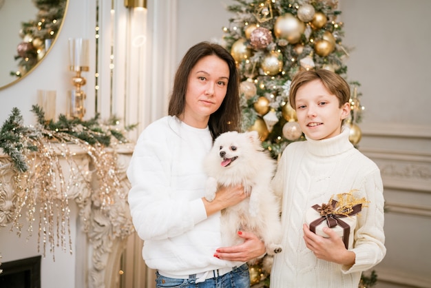 Schöne lächelnde Mutter und Sohn, die sich zu Hause mit einem glücklichen weißen Hund für den Weihnachtsurlaub umarmen