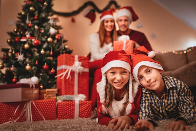 Schöne lächelnde Schwester und Bruder, die sich in der Weihnachtszeit zu Hause amüsieren. Sie tragen Weihnachtsmützen und schauen in die Kamera. Selektiver Fokus. Fokus auf Vordergrund, auf Kinder.