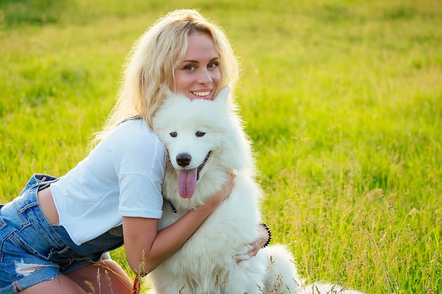 Schöne lockige blonde lächelnde glückliche junge Frau in Denim-Shorts, die spielt, umarmt einen weißen, flauschigen, süßen Samojeden-Hund im Sommerpark-Sonnenuntergang-Strahlen-Feldhintergrund. Haustier und Gastgeberin