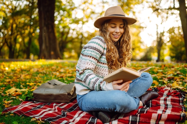 Schöne lockige Frau mit Hut auf Matte mit Buch im Herbstpark Entspannung, Einsamkeit mit der Natur
