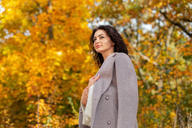 Schöne lockige Herbstmodefrau in einem stylischen Mantel mit Pullover geht in einem goldenen Herbstpark spazieren