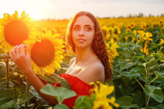 Schöne lockige junge Frau im Sonnenblumenfeld, die Sonnenblumenblume in der Hand hält Porträt des jungen W...