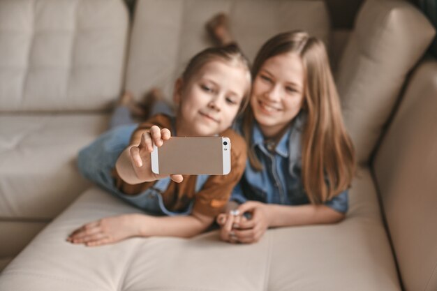 Schöne Mädchen machen Selfies auf ihrem Handy, während sie auf dem Sofa liegen. Schwestern machen eine Pause von den Hausaufgaben und machen während der Quarantäne Fotos