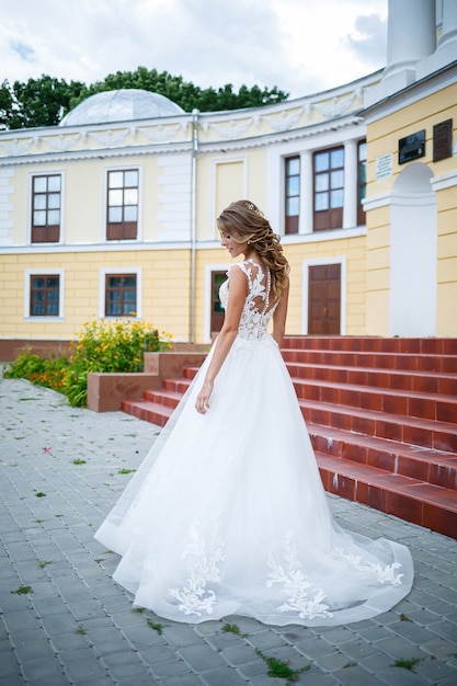 Schöne Mädchenbraut in einem weißen Kleid mit einem Zug, der an ihrem Hochzeitstag auf dem Hintergrund eines großen Hauses mit Säulen geht