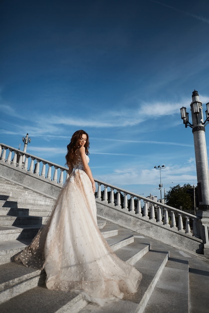 Schöne Modellfrau mit perfektem Körper im Luxushochzeitskleid steht mit zurück auf der Treppe und posiert mit blauem Himmel am Hintergrund