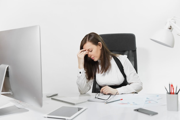 Schöne müde verwirrte und gestresste braunhaarige Geschäftsfrau in Anzug und Brille, die am Schreibtisch sitzt und an einem modernen Computer mit Dokumenten und Monitor im hellen Büro arbeitet working
