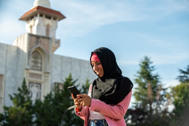 Schöne muslimische Frau, die einen Hijab mit Technologie mit Moscheehintergrund trägt. Horizontale Ansicht der arabischen Frau mit Telefon im Freien. Technologie und Lebensstil der muslimischen Frauen.