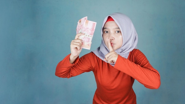 Schöne muslimische Frau, die Indonesien-Rupiah-Geld hält und darum bittet, ruhig zu sein, mit dem Finger auf den Lippen Stille und geheimes Konzept