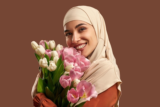 Schöne muslimische Frau in beigem Hijab mit Tulpenstrauß lächelnd Schönheitsfrühlingskonzept