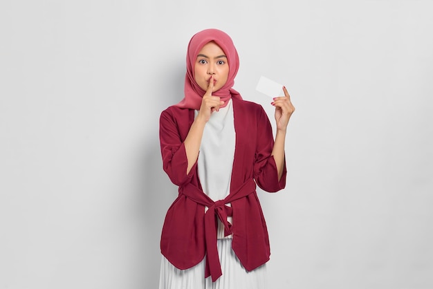 Schöne muslimische Frau in Freizeithemd und Hijab, die Kreditkarte zeigt, Finger, die Lippen bedecken, macht Schweigegeste isoliert auf weißem Hintergrund. Menschen religiöses Lebensstilkonzept