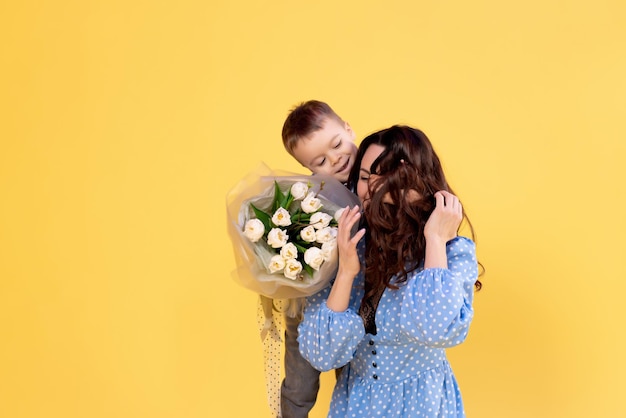 Schöne Mutter mit einem kleinen Jungen auf gelbem Hintergrund Liebevolle fürsorgliche Mutter spielt mit ihrem Sohn im Vorschulalter Ein Junge mit einem großen Blumenstrauß Entwicklung des Kindes