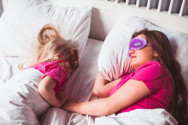Schöne Mutter und ihre kleine Tochter schlafen im Bett mit Schlafmasken Sie stehen sich gegenüber und halten sich an den Händen