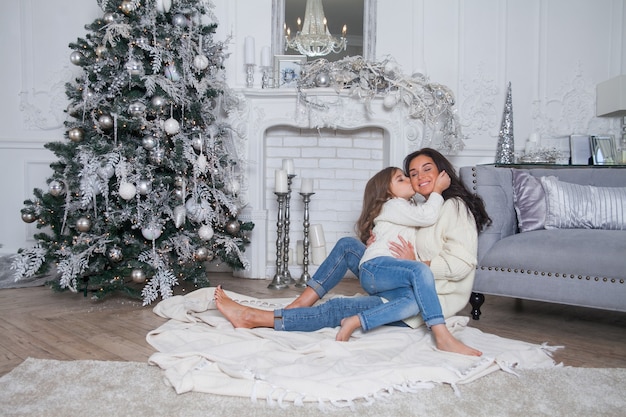 Schöne Mutter und süße Tochter in weißen gemütlichen Pullovern haben Spaß und Umarmung unter einem geschmückten Weihnachtsbaum. Festliches Zuhause klassisches Interieur mit Kamin und grauem Sofa.