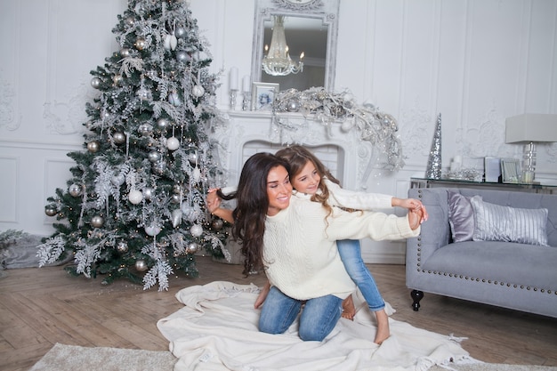 Schöne Mutter und süße Tochter in weißen gemütlichen Pullovern haben Spaß und Umarmung unter einem geschmückten Weihnachtsbaum. Festliches Zuhause klassisches Interieur mit Kamin und grauem Sofa.