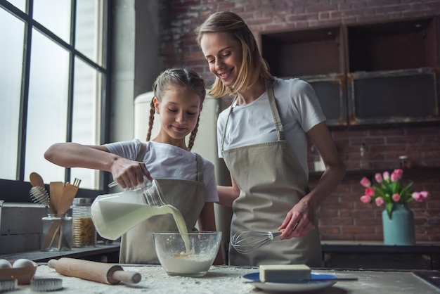 Schöne Mutter und Tochter in Schürzen fügen Milch zu Mehl hinzu und lächeln, während sie zu Hause Kekse backen