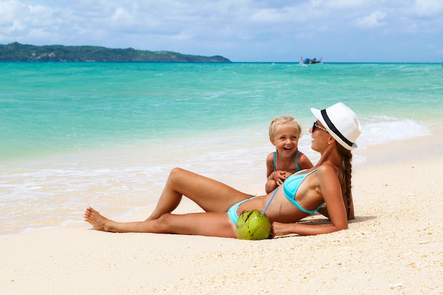 Schöne Mutter und Tochter liegen im Badeanzug am Strand mit weißem Sand und Lachen