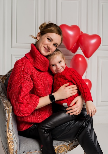 Schöne Mutter und Tochter sitzen und umarmen in einem Stuhl auf einem weißen Hintergrund mit roten Luftballons