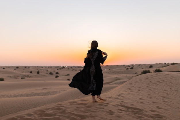 Schöne mysteriöse Frau im traditionellen arabischen schwarzen langen Kleid steht bei Sonnenuntergang in der Wüste