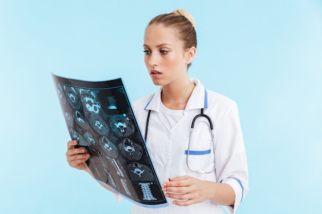 Schöne nachdenkliche blonde Ärztin in Uniform, die isoliert über blauer Wand steht und Röntgen analysiert
