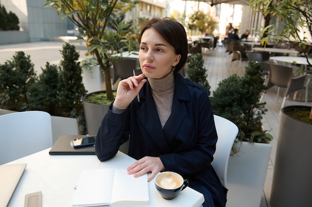 Schöne nachdenkliche Frau, die zur Seite schaut, einen Stift hält und Projekte plant, in ein Notizbuch schreibt, im Freien arbeitet, mit einem Laptop, Smartphone und einer Tasse Kaffee an einem Tisch sitzt