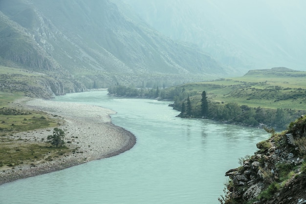 Schöne neblige Berglandschaft mit breitem Gebirgsfluss. Dunkelgrüne düstere Landschaft mit großem Gebirgsfluss im Nebel. Dunkler atmosphärischer Blick auf den großen Fluss zwischen den großen Bergen bei Regenwetter.