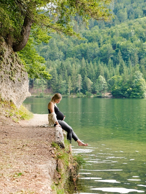 Schöne, niedliche schwangere Frau sitzt nahe einem Bergsee in einem Wald am Ufer