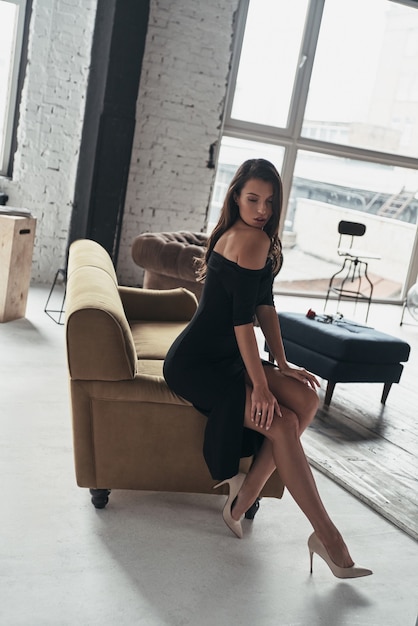 Schöne Perfektion. Blick von oben auf eine attraktive junge Frau im eleganten schwarzen Kleid mit tiefem Schlitz, die beim Sitzen auf dem Sofa posiert