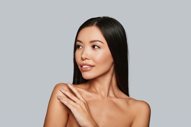 Schöne Perfektion. Schöne junge asiatische Frau, die beim Stehen gegen grauen Hintergrund lächelt