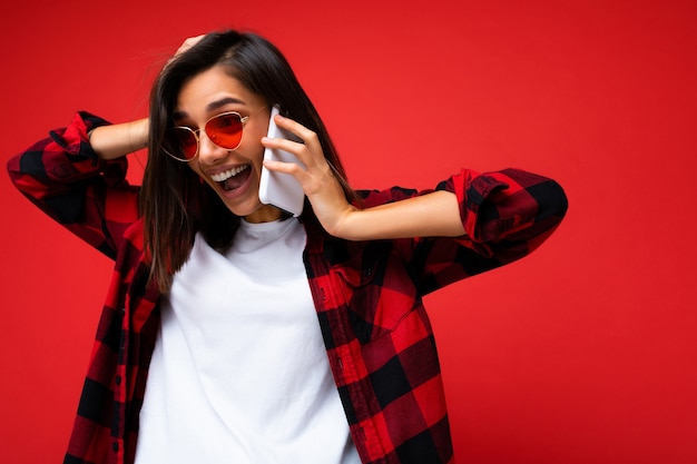 Schöne positive schockierte erstaunte junge brünette Frau mit stilvollem rotem Hemd, weißem T-Shirt und roter Sonnenbrille einzeln auf rotem Hintergrund, die mit dem Handy spricht und Spaß hat. Platz kopieren