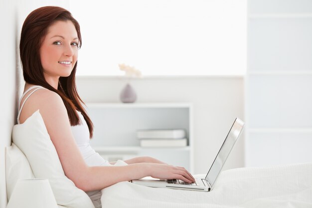 Schöne redhaired Frau, die mit ihrem Laptop beim Sitzen auf ihrem Bett sich entspannt