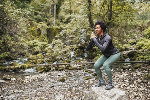 Foto schöne reife afroamerikanische läuferin, die kniebeugenübungen in der nähe des flusses in der natur macht.