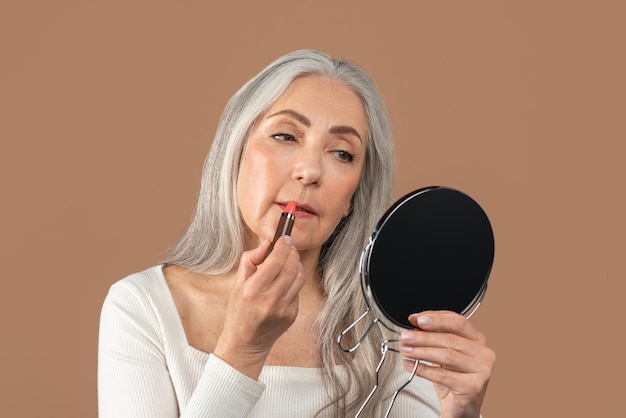 Schöne reife Dame, die Lippenstift vor dem Spiegel aufträgt und sich im braunen Studio um sich selbst kümmert