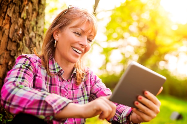 Schöne reife Frau im Park mit einem digitalen Tablet