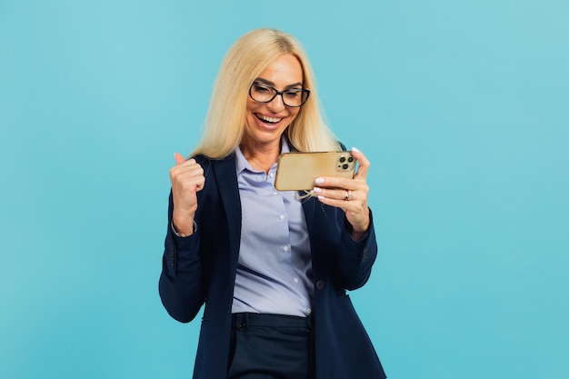 Schöne reife Frau in Business-Anzug und Brille hält am Telefon und zeigt sich gut vor isoliertem blauem Hintergrund