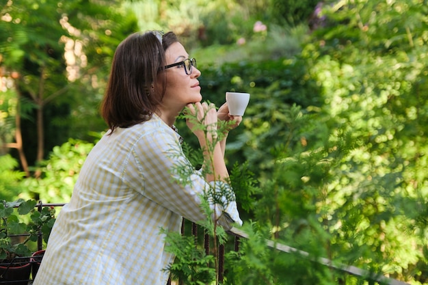 Schöne reife Frau in der Hauskleidung auf offenem Balkon mit Tasse Kaffee