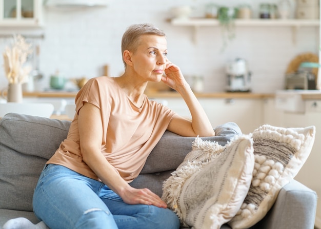 Schöne reife Frau in Freizeitkleidung, die von etwas träumt, während sie sich zu Hause auf dem Sofa entspannt