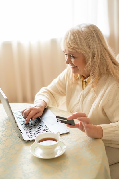 Schöne reife Frau kauft im Internet mit einer Kreditkarte ein, während sie am Tisch sitzt und eine Tasse Tee trinkt