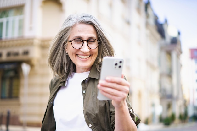 Schöne reife Frau mit grauen Haaren, die auf ein weißes Smartphone schaut und durch die Straßen einer alten europäischen Stadt spaziert. Reife Frau liest Textnachrichten oder beantwortet Videoanrufe im Stehen oder Gehen im Freien