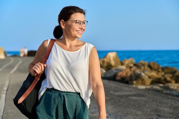 Schöne reife Frau mit Rucksack, die draußen am Reiseziel am Meer spaziert