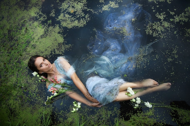 Schöne romantische Frau der Kunst liegt im Sumpf im blauen langen Kleid mit Blumen. Porträt Brunette im transparenten Kleid in Wassersumpf-Schlamm-Entengrütze. Buchcover