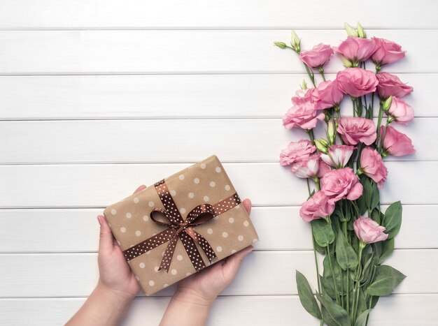 Schöne rosa Eustoma Blumen und Frauenhände hält handgemachte Geschenkbox auf weißem hölzernem Hintergrund. Speicherplatz kopieren, Draufsicht,