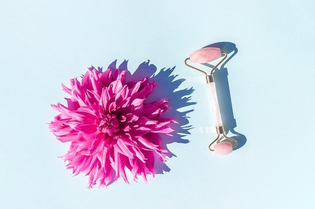 Schöne rosafarbene Blume und Kristall-Rosenquarz-Gesichtsrolle auf blauem Hintergrund Gesichts-Antiage-Massage Konzept Jugendschönheit und Selbstpflege