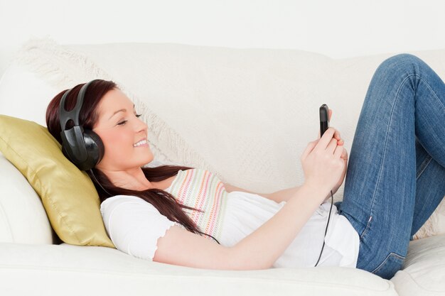 Schöne rothaarige Frau, die Musik mit Kopfhörern beim Lügen auf einem Sofa hört