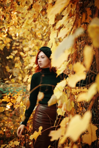 Schöne rothaarige Frau im Herbstparkmädchen im schwarzen Barettstrickpullover und im Lederrockstil...