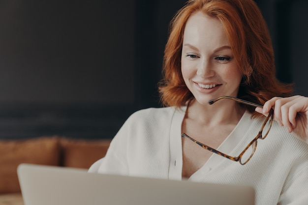Schöne rothaarige Frau konzentriert in Laptop-Display, lächelt positiv, hält optische Brille, posiert drinnen, nutzt moderne Technologien für die Online-Kommunikation, sieht Webinar, liest Nachrichten