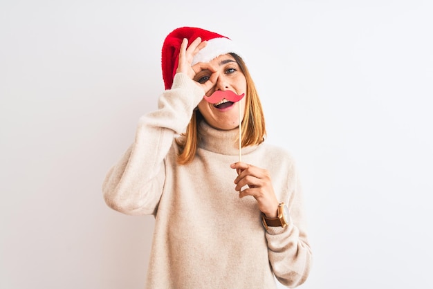 Schöne rothaarige Frau mit Weihnachtsmütze und vorgetäuschtem Schnurrbart vor isoliertem Hintergrund mit glücklichem Gesicht, lächelnd und mit der Hand auf dem Auge, die durch die Finger schaut, ein „OK“-Zeichen machend