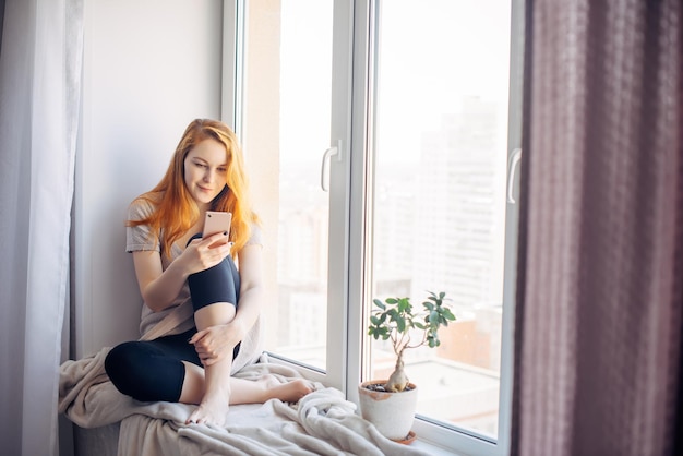 Foto schöne rothaarige frau sitzt auf der fensterbank in einer gemütlichen stadtwohnung junges mädchen verbringt ihre freizeit mit dem smartphone